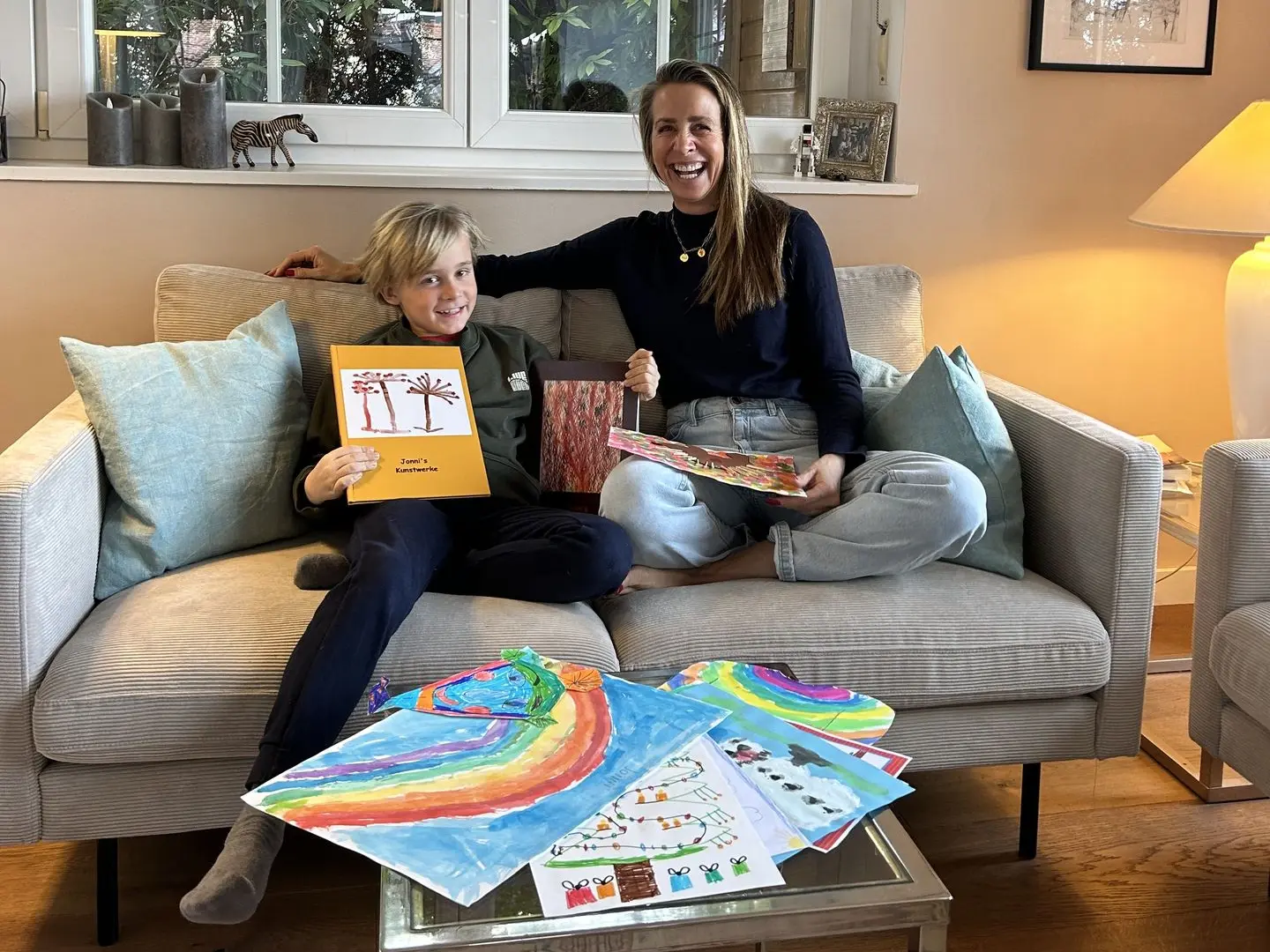 Jonni`s KIDSartbook Glücklicher 10 jähriger Bub sitzt glücklich mit lachender Mama am Sofa, Kinderzeichnungen liegen am Tisch davor und der Bub hat sein eigenes KIDSartbook, ein Buch mit seinen eingescannten Bildern, in der hat und schaut sehr stolz auf seine Kunstwerke.